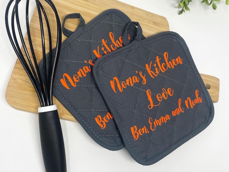 May include: Two gray quilted pot holders with orange text. The text on one pot holder reads "Nona's Kitchen Love Ben, Emma and Noah". The text on the other pot holder reads "Nona's Kitchen".