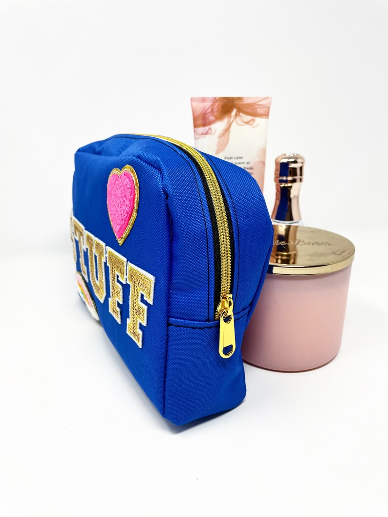 Op de afbeelding: Een blauwe toilettas met een gouden rits en een gouden geborduurde patch met de tekst "STUFF" en een roze hartpatch.