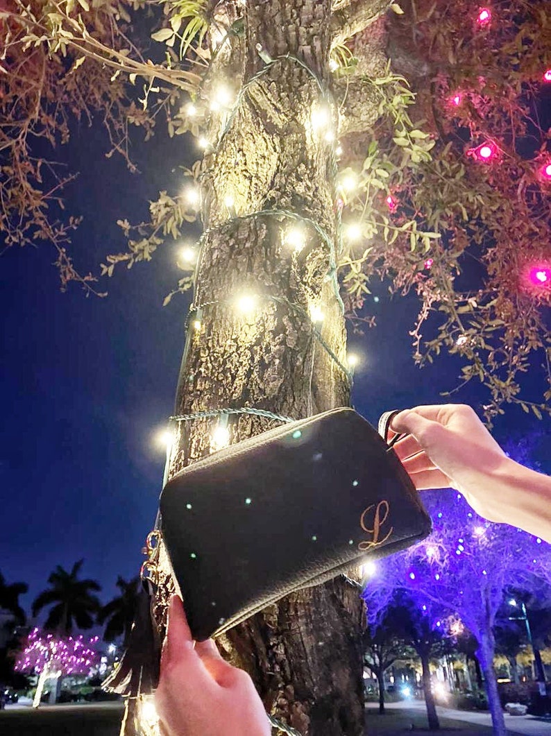 May include: A black clutch bag with a gold tassel and the letter "L" embroidered on the front. The bag is held in front of a tree with white lights wrapped around it.