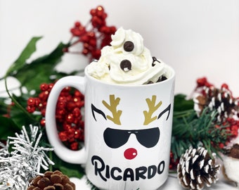 May include: White ceramic mug with a reindeer design, wearing sunglasses and a red nose. The mug has the name "RICARDO" printed on it. The mug is filled with whipped cream and chocolate chips.