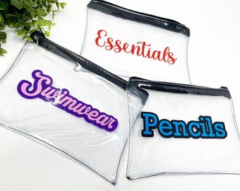 Puede incluir: Tres bolsas transparentes de plástico con cremallera y ribete negro. Las bolsas están etiquetadas con "Essentials" en rojo, "Swimwear" en morado y rosa, y "Pencils" en azul.