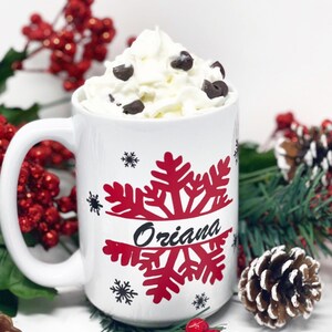 May include: White ceramic mug with a red snowflake design and the name "Oriana" written in black. The mug is filled with whipped cream and chocolate chips and is surrounded by red berries and pine branches.