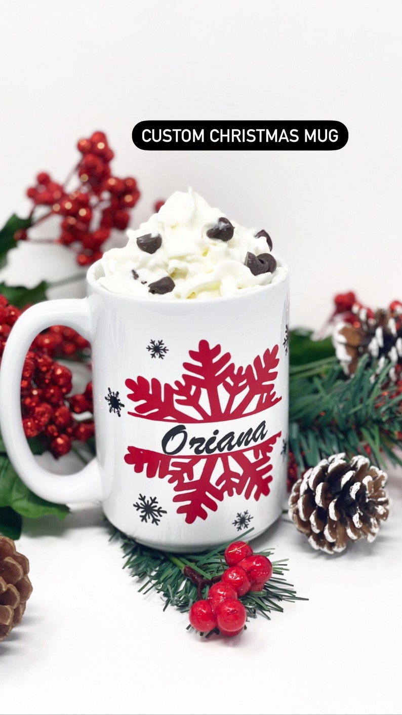 May include: White ceramic mug with a red snowflake design and the name "Oriana" written in black. The mug is filled with whipped cream and chocolate chips and is surrounded by red berries and pine branches.
