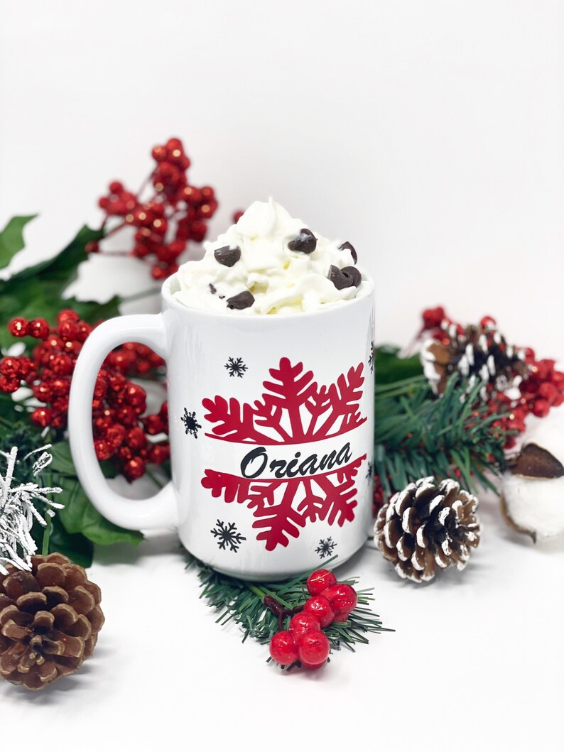 May include: White ceramic mug with a red snowflake design and the name "Oriana" written in black. The mug is filled with whipped cream and chocolate chips. The mug is surrounded by green pine branches and red berries.