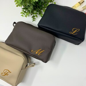 May include: Three toiletry bags in different colors with personalized initials embroidered on them. The bags are beige, brown, and black.