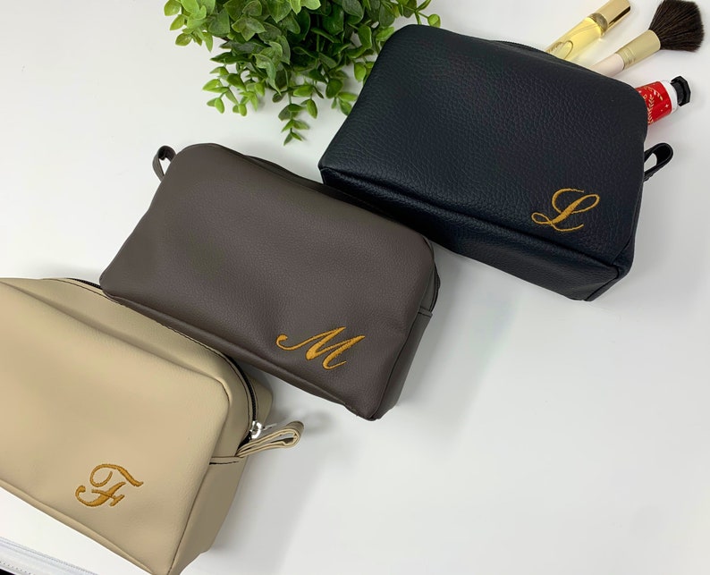 May include: Three toiletry bags in different colors with personalized initials embroidered on them. The bags are beige, brown, and black.
