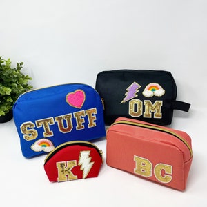 Op de afbeelding: Vier verschillende toilettassen met gepersonaliseerde letters en patches. De tassen zijn in blauw, zwart, koraal en rood. De patches omvatten een hart, een bliksem, een regenboog en de letters "OM".