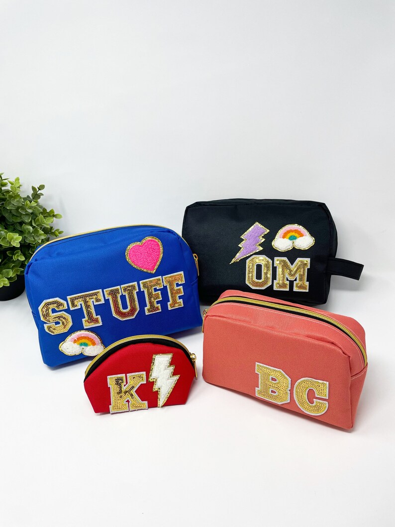 Op de afbeelding: Vier verschillende toilettassen met gepersonaliseerde letters en patches. De tassen zijn in blauw, zwart, koraal en rood. De patches omvatten een hart, een bliksem, een regenboog en de letters "OM".