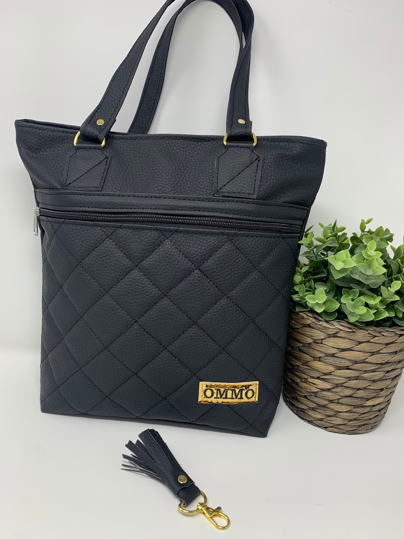 May include: Black quilted tote bag with gold hardware and a zippered pocket. The bag has a gold label with the text "OMMO" on it. A black tassel keychain with a gold clasp is included.