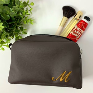 May include: A brown faux leather zippered travel makeup bag with a gold embroidered letter M.