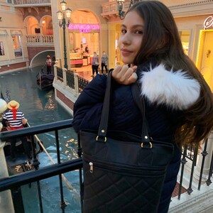 May include: A young person wearing a black puffer jacket with a white fur trim and a black quilted tote bag. The person is standing in front of a building with a canal in the foreground.