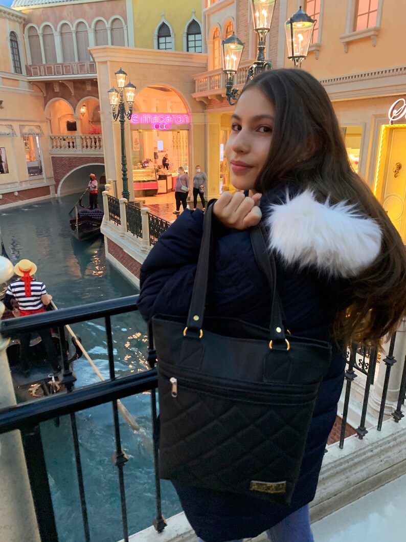 May include: A young person wearing a black puffer jacket with a white fur trim and a black quilted tote bag. The person is standing in front of a building with a canal in the foreground.