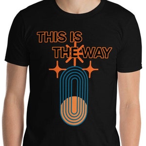 K&ouml;nnte beinhalten: Schwarzes T-Shirt mit dem Text "This is the way" in orangefarbener und schwarzer Schrift. Unter dem Text befindet sich ein stilisiertes orangefarbenes und blaues Symbol.