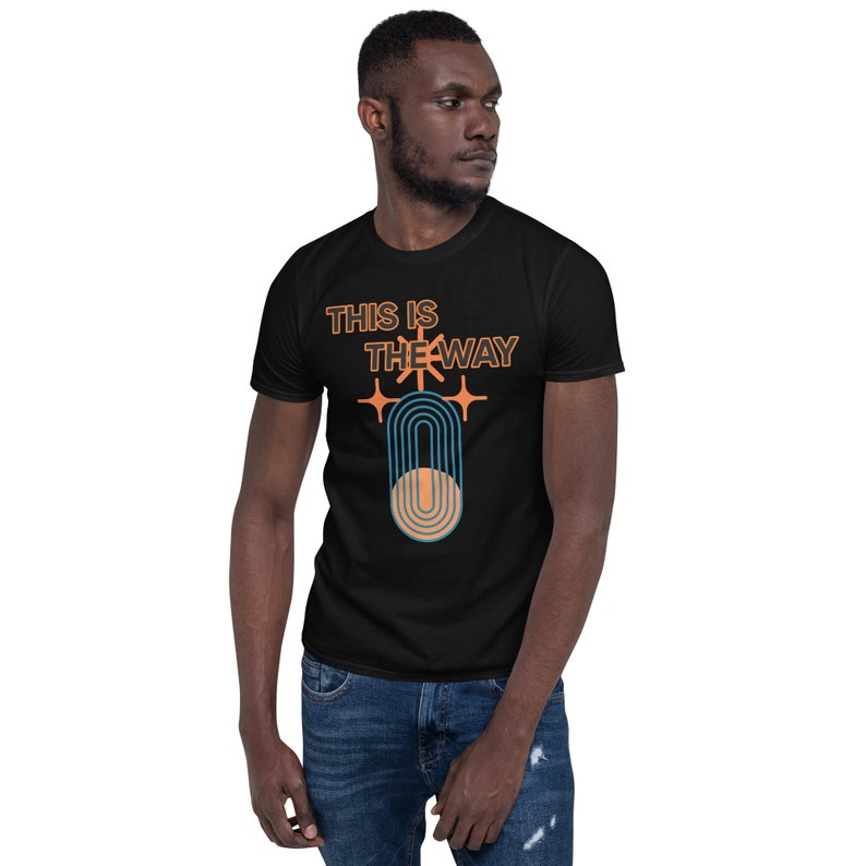 K&ouml;nnte beinhalten: Schwarzes T-Shirt mit dem Text "This is the way" in orangefarbener und blauer Schrift. Das Shirt zeigt au&szlig;erdem eine stilisierte Grafik eines Halbmondes mit einem Regenbogenbogen in Orange und Blau.
