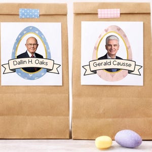 May include: Brown paper bags with white labels featuring portraits inside egg-shaped frames. The labels display the names "Dallin H. Oaks" and "Gerald Causse" on banners. The bags are decorated with washi tape and surrounded by colorful Easter eggs.