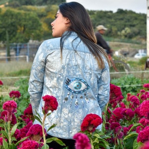 May include: A woman wearing a light blue denim jacket with a large, colorful eye design on the back. The jacket is decorated with silver sequins and beads. The woman is standing in a field of pink flowers.