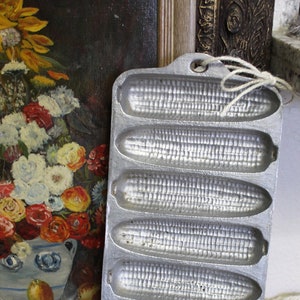 May include: A silver metal cornbread pan with six individual molds shaped like ears of corn. The pan has a handle on the top.