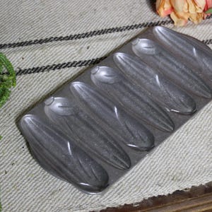 May include: A rectangular cast iron baking pan featuring five distinct, elongated, corn-shaped molds. The metallic gray surface has a textured finish, and the mold is designed to create five individual corn-shaped baked goods in a single batch.