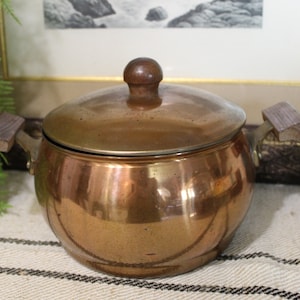 May include: A copper pot with a wooden lid and handles. The pot is round and has a shiny finish.