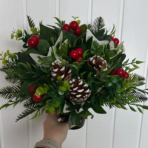 May include: A Christmas-themed arrangement with a mix of green foliage, red berries, and pine cones. The arrangement features various types of leaves and branches, creating a full and textured appearance. The pine cones are brown, and the berries are a vibrant red.