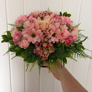 May include: A floral arrangement featuring a central peach-coloured dahlia surrounded by various shades of pink roses, daisies, and small hydrangea clusters. The bouquet is accented with vibrant green foliage and delicate fern-like leaves.