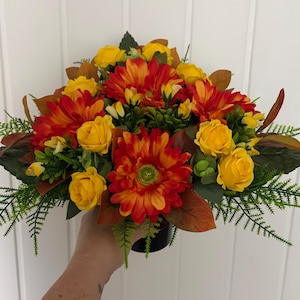 May include: A floral arrangement featuring vibrant orange and yellow gerbera daisies, small yellow roses, and green foliage. The bouquet includes dark brown leaves and feathery green fern-like sprigs, all gathered together in a dark circular base.