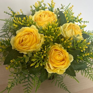May include: A close-up of a floral arrangement with five yellow roses as the focal point. The roses are surrounded by green leaves, including fern and eucalyptus. Small yellow buds add to the arrangement's texture. A bright and colourful display.