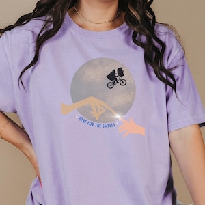 May include: A purple t-shirt with a graphic of a silhouette of two figures riding a bicycle in front of a moon with the text "Here for the Smells".