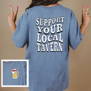 May include: A blue t-shirt with the text "SUPPORT YOUR LOCAL TAVERN" printed on the back. The front of the shirt has a small graphic of a beer mug with foam.