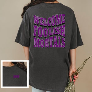 May include: A dark gray oversized t-shirt with the text "WELCOME FOOLISH MORTALS" printed in purple on the back. The shirt has a relaxed fit and a crew neck.