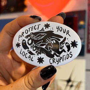 Op de afbeelding: Een ovale witte pinback-button met een zwarte grafische illustratie van diverse wezens, waaronder een jackalope en geesten, omringd door de tekst "PROTECT YOUR LOCAL CRYPTIDS" en kleine stervormige accenten in zwarte inkt.