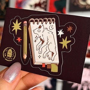 Può includere: Un adesivo marrone scuro con un disegno di un lupo in un taccuino, una matita e stelle. L'adesivo ha anche il testo "Mango Milk Studio". Lo sfondo include varie stampe artistiche.