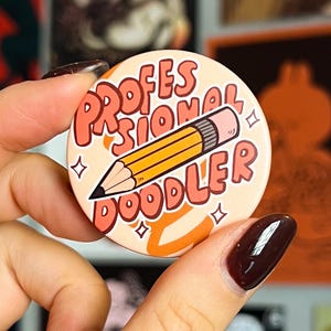 Könnte beinhalten: Ein runder Button mit dem Text "PROFESSIONAL DOODLER" und einer gelben Bleistiftillustration. Der Button hat einen pfirsichfarbenen Hintergrund mit orangefarbenen Akzenten. Er wird in einer Hand mit dunklem Nagellack gehalten.