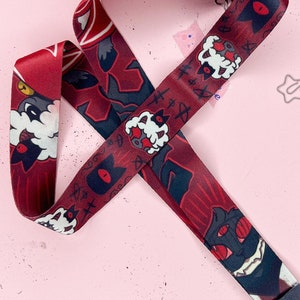 May include: A burgundy lanyard with a repeating pattern of cartoon-style characters with white and black accents. The lanyard has a dark gray leather-like end piece with a silver-colored metal clasp. The background is a light pink color.