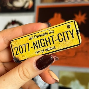 Könnte beinhalten: Ein gelber Magnet in Form eines Nummernschilds mit dem Text "2077-NIGHT-CITY CITY OF DREAMS" und "Del Coronado Bay". Der Magnet hat einen Used-Look. Im Hintergrund befinden sich verschiedene Kunstdrucke.