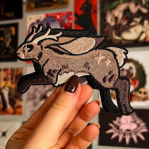 Op de afbeelding: Geborduurde patch met een bruine jackalope met geweien, rennend naar rechts. De patch heeft een gedetailleerd ontwerp met verschillende bruin- en beigetinten, met een zwarte omtrek. De achtergrond is een verzameling kunstprints.