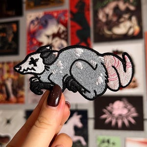 Op de afbeelding: Geborduurde patch met een gestileerd opossum-ontwerp. De patch heeft een grijze body met witte accenten, zwarte poten en een zwart-wit gezicht. De staart van de opossum is roze en gestreept. De patch wordt tegen een achtergrond van diverse kunstdrukken gehouden.