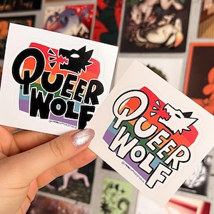 May include: Two white square stickers with the text "Queer Wolf" in black and rainbow colors, featuring a wolf head graphic. The stickers are held in front of a wall covered in various art prints.