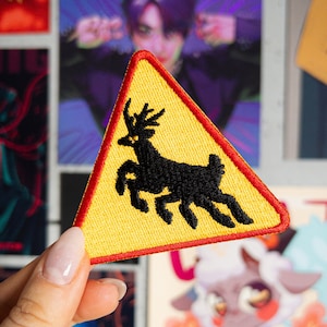 Könnte beinhalten: Ein gelbes dreieckiges Warnschild mit rotem Rand. Das Schild hat eine schwarze Silhouette eines Rehs darin. Das Schild wird von einer Hand gehalten.