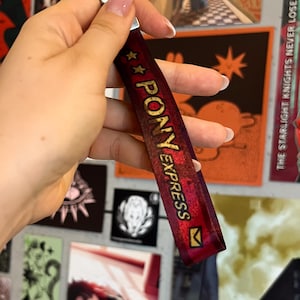 Peut inclure: Un cordon de cou bordeaux avec les mots "PONY EXPRESS" en lettres dorées et un fermoir argenté. Le cordon porte des étoiles dorées et un petit graphisme d'enveloppe. L'arrière-plan présente diverses impressions d'art.