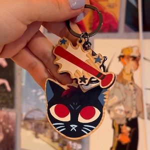 May include: Wooden keychain with a black cat face and red eyes, plus a baseball bat design. The keychain is attached to a metal ring. The cat has blue accents and white whiskers. The baseball bat design has red and white accents.