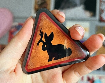 May include: A triangular, orange and black decorative pin featuring a black silhouette of a rabbit holding a branch. The pin has a black border and is held in a hand.