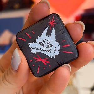 May include: A black square pin featuring a white wolf design with red accents. The pin has a textured surface and is held in a hand against a backdrop of posters and a heart-shaped light.