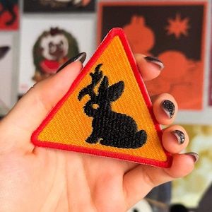 Könnte beinhalten: Ein gelbes Dreieck-Warnschild mit rotem Rand, das eine schwarze Silhouette eines Kaninchens mit Geweih zeigt. Das Schild ist aus Stoff und hat ein gesticktes Design.