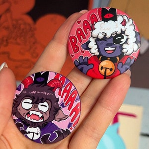 Könnte beinhalten: Zwei runde rosa und violette Knöpfe mit Comicfiguren. Der erste Knopf zeigt ein lila Schaf mit einem roten Halsband und einer Glocke, mit dem Text "BAAA!" in rosa. Der zweite Knopf zeigt eine lila Ziege mit Hörnern, mit dem Text "AAAA!" in rosa.