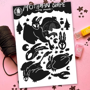 May include: A sheet of black vinyl stickers featuring various Mothman shapes, stars, and trees. The sheet includes the text "Mothman Shape - Vinyl Stickers" and the Mango Milk Studio logo. A roll of black twine and a pink stapler are visible.