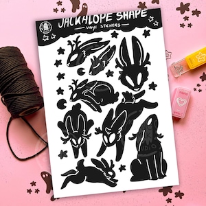 Op de afbeelding: Een vel zwarte vinylstickers met jackalope-vormen en sterren. De stickers staan op een witte achtergrond met bovenaan de tekst "JACKALOPE SHAPE VINYL STICKERS". Een rol bruin touw en twee kleine roze en gele voorwerpen staan op de achtergrond.