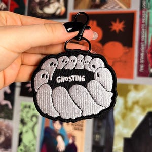 Op de afbeelding: Zwarte geborduurde patch met een cirkel van witte geesten en het woord "Ghosting" in het midden. De patch is bevestigd aan een zwarte metalen sleutelhanger.