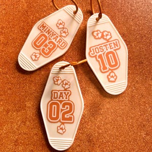 May include: Three off-white motel-style keychains with orange text and paw prints. The keychains read "MINYARD 03", "JOSTEN 10", and "DAY 02". Each keychain has a gold-colored metal ring attached at the top.