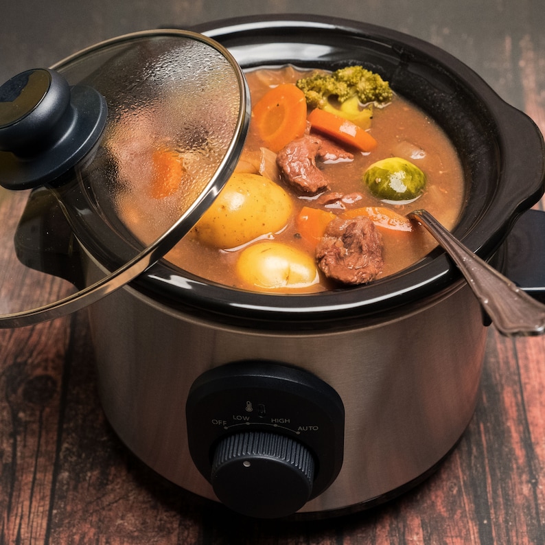 May include: A silver slow cooker with a glass lid, filled with a stew of beef, potatoes, carrots, and Brussels sprouts. The slow cooker has a dial with settings for off, low, high, and auto.